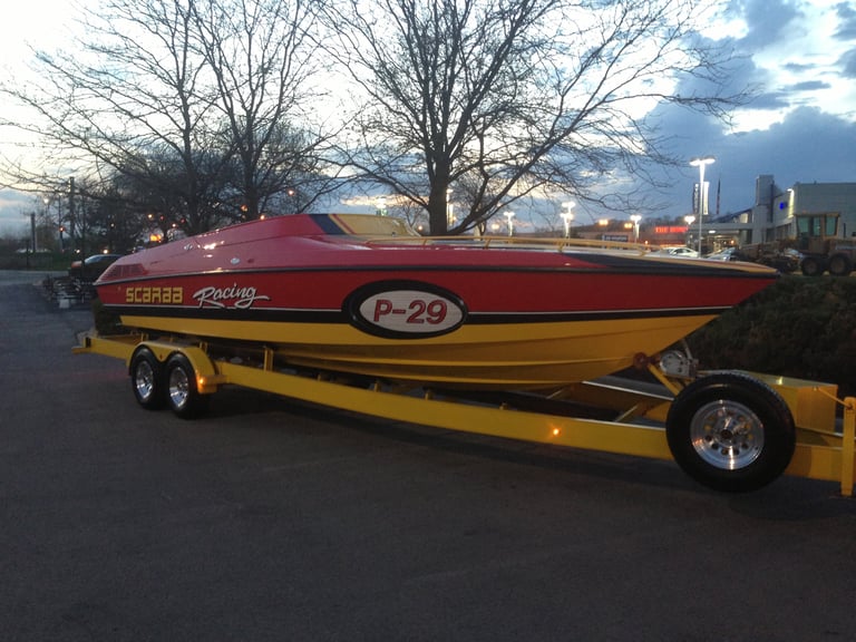 Red and yellow Scarab Racing P-29 on its dual-axle trailer at twilight, parked in a lot with bare trees and a city skyline behind