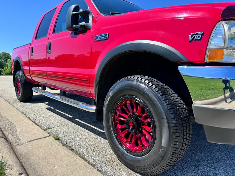 Side profile of the red 2003 Ford F-250 Super Duty on the street, with the chrome side step running the length of the cab, the F-250 and Triton V10 badges on the front fender, and the custom red fuel forged alloy wheel with Mickey Thompson all-terrain tire taking up the foreground
