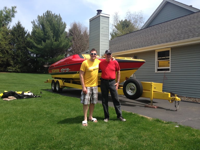 Two men in yellow and red shirts standing on a lawn in front of a red and yellow P-29 Scarab Racing boat on a yellow trailer