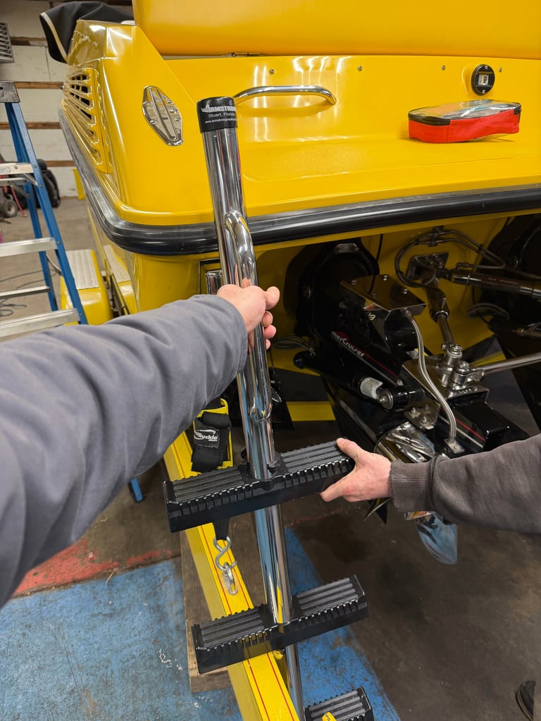Two sets of hands test-fitting the new chrome 4-step Ameristep boarding ladder against the transom of the yellow P-29, with the Mercury outdrive and swim platform hardware below