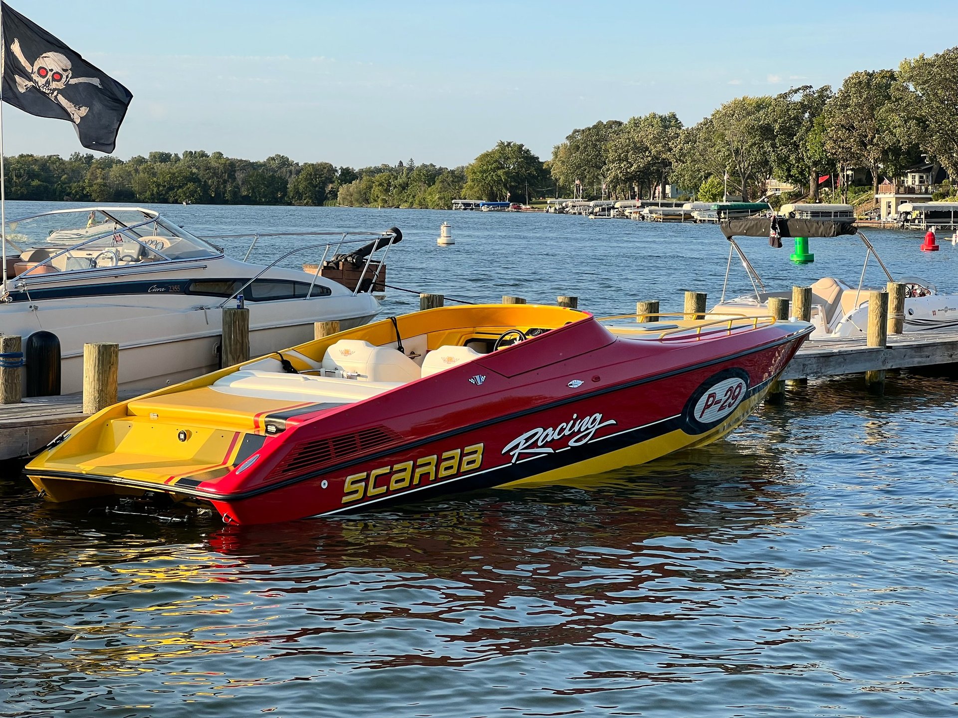 A red and yellow speedboat docked at a wooden pier on a blue lake with residential shoreline and trees in the background