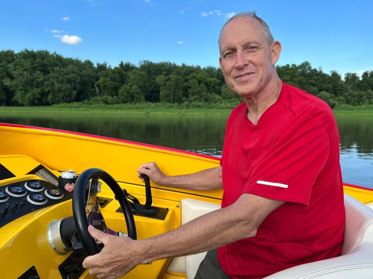 Man in a red t-shirt smiling at the helm of the yellow P-29, throttle in his right hand, looking out over a river lined with green trees under a clear blue sky