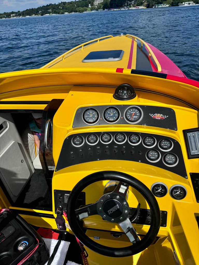 Yellow and pink speedboat cockpit with steering wheel and multiple gauges, docked on blue water with waterfront homes visible
