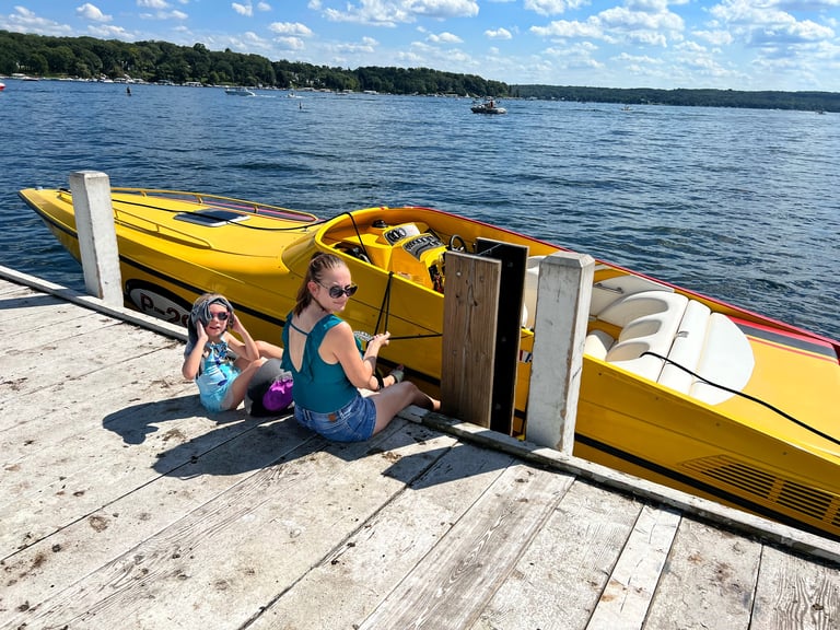 Two children sitting on a dock next to a yellow and orange small boat with P-29 markings on a sunny lake