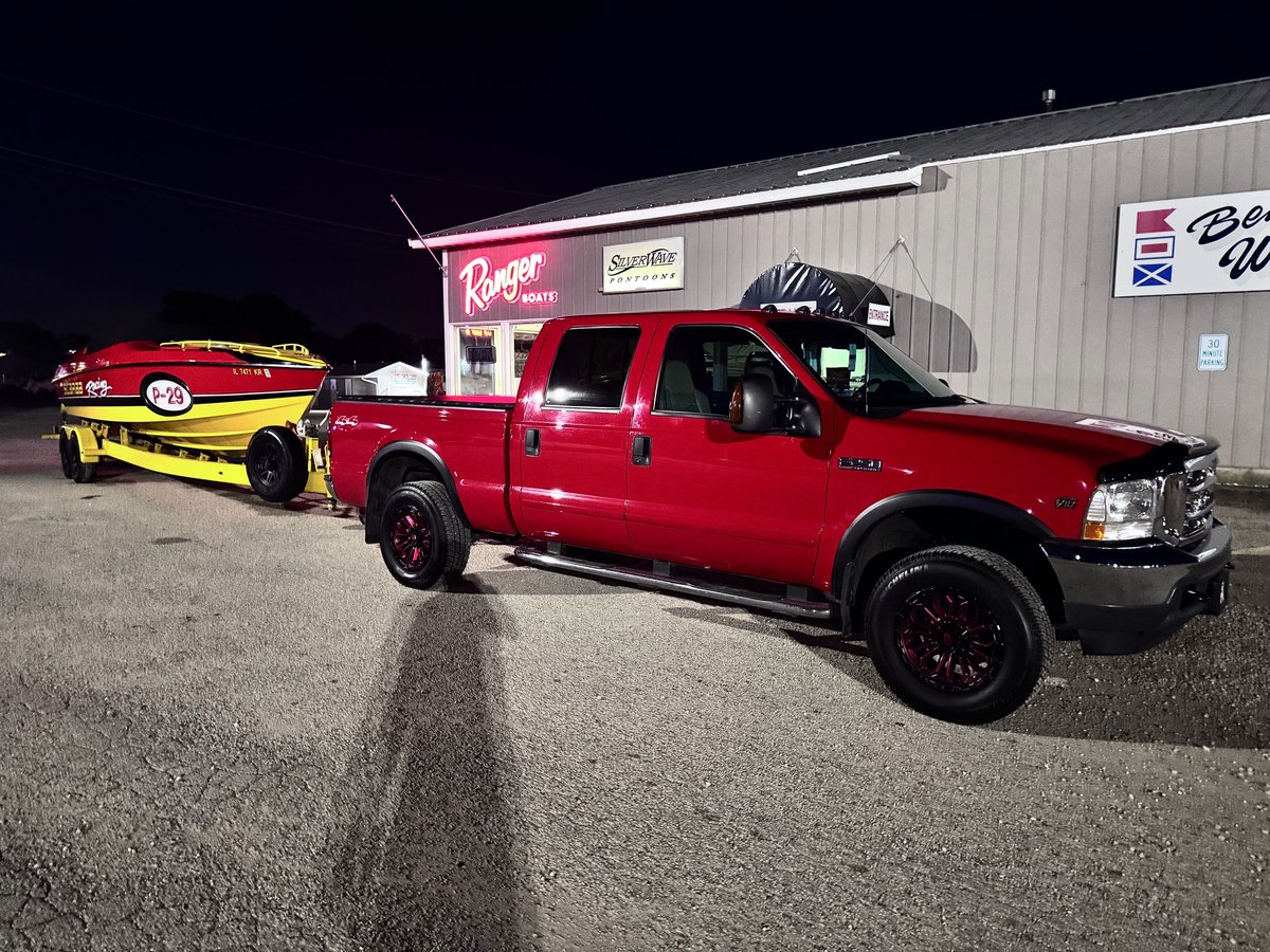 Red F-250 with red 4x4 off-road wheels hitched to the yellow P-29 on its trailer at night, lit by the neon Ranger Boats and Silverwave Pontoons signs outside a vintage shop