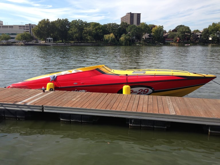 Red and yellow speedboat docked on wooden pier with city buildings and trees along river in background