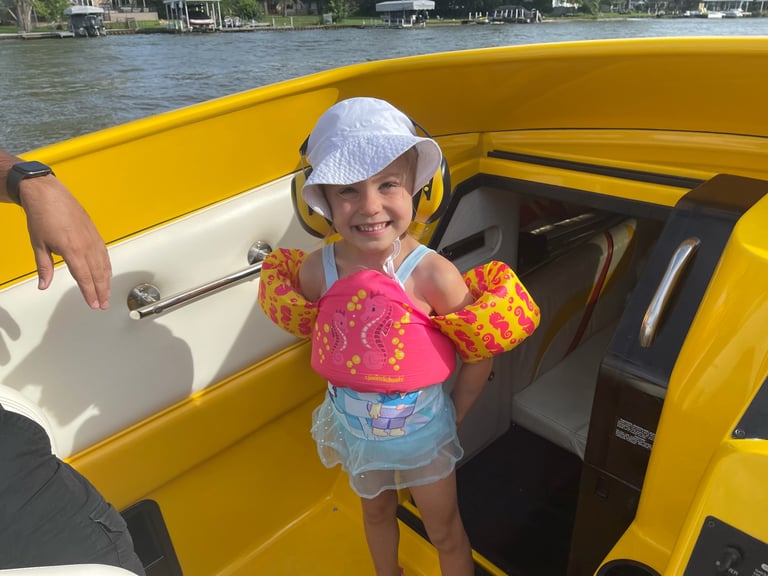 Child wearing white cap and pink water wings smiling in yellow speedboat on water