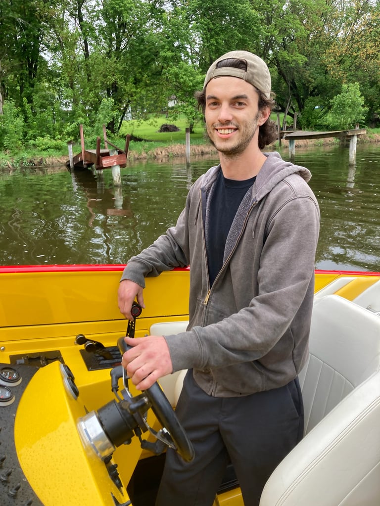 Man in gray hoodie and cap holding fishing rod on yellow boat in river with trees in background