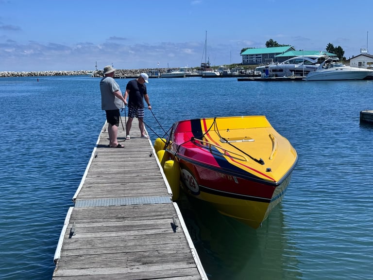 Two people standing on a wooden dock beside a red and yellow speedboat tied up at a marina