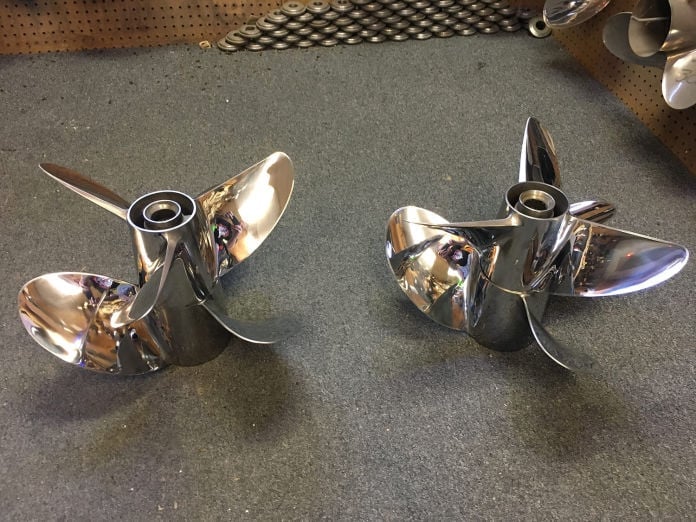 Two polished stainless steel four-blade Mercury Racing propellers displayed on a dark counter, with extra props visible on a pegboard wall behind