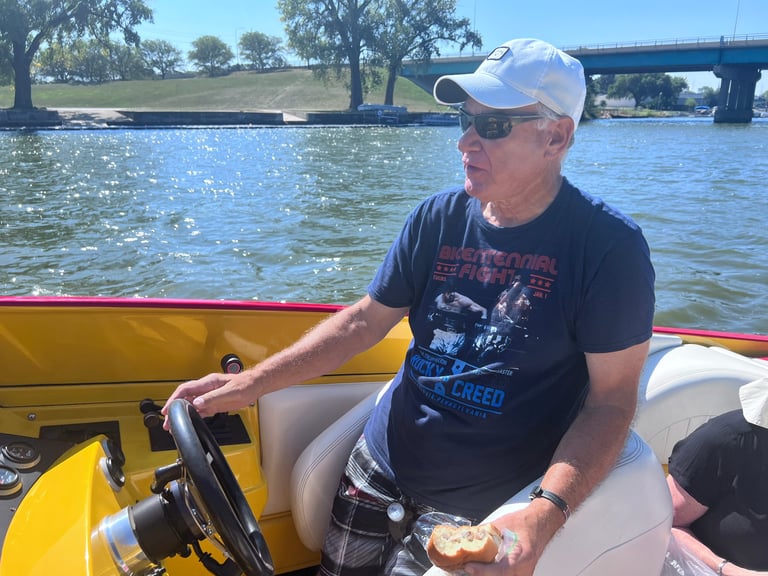 Man in white cap and black shirt sitting in yellow and white speedboat on river with bridge in background