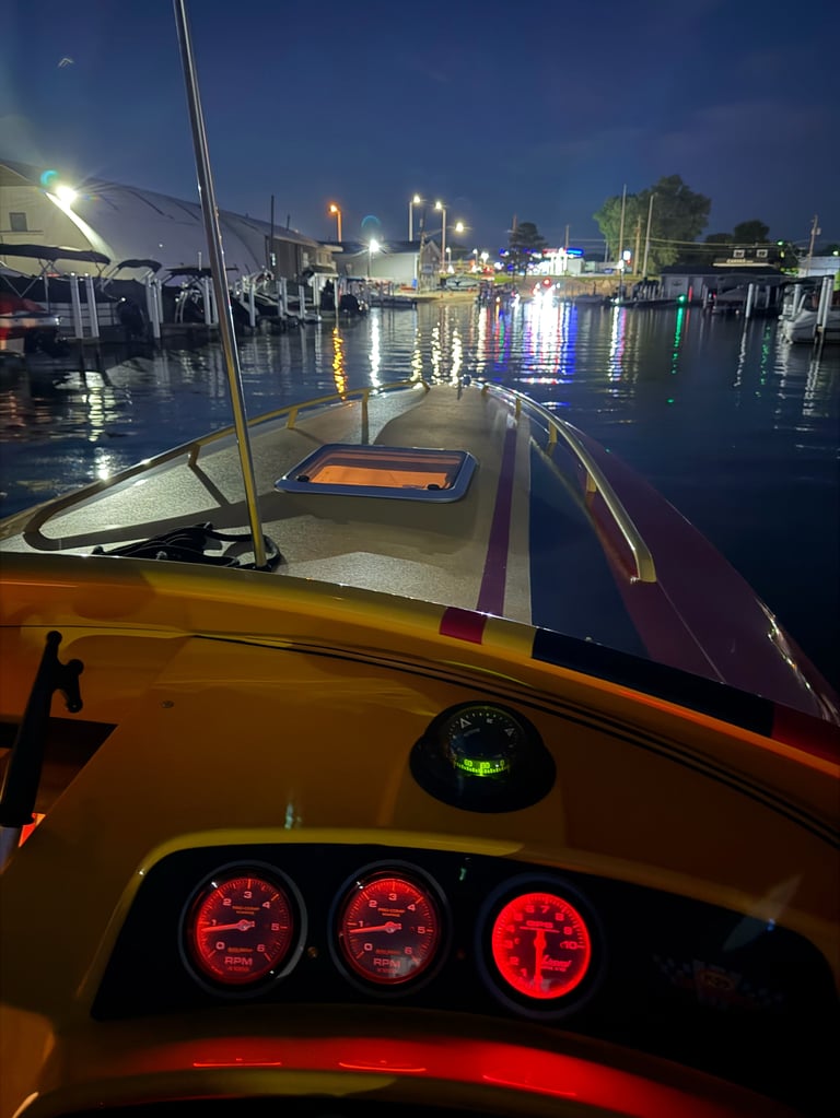 Night view from the helm of the P-29, red-lit Livorsi RPM gauges and compass glowing against the dark, with the yellow bow and deck hatch ahead and marina lights reflecting off calm water