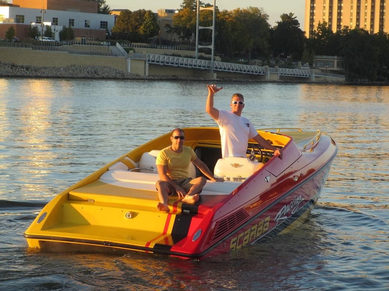 Two men aboard the yellow and red Scarab P-29 on a river at golden hour, one waving, with city buildings and trees along the bank behind them