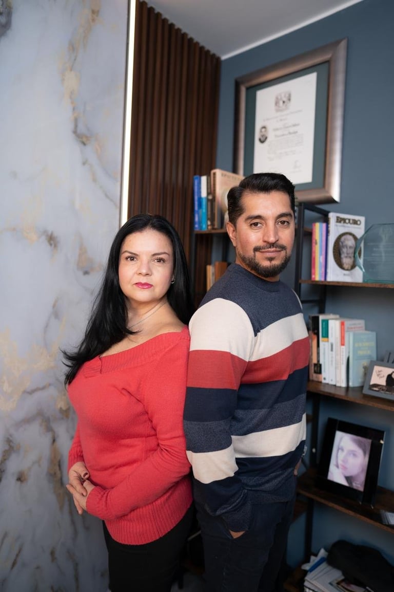 Mujer embarazada con suéter rojo y hombre con suéter a rayas posando juntos en consultorio moderno con paredes azules y estantes con libros