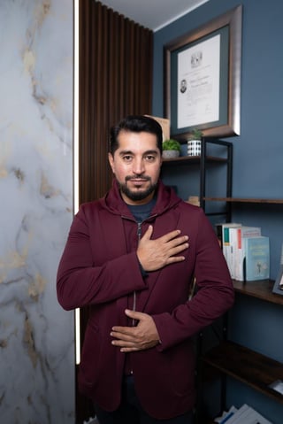 Psic. Walberto Tenorio, especialista en Terapia Breve Basada en Soluciones, con mano en el pecho frente a estantería azul y certificado enmarcado