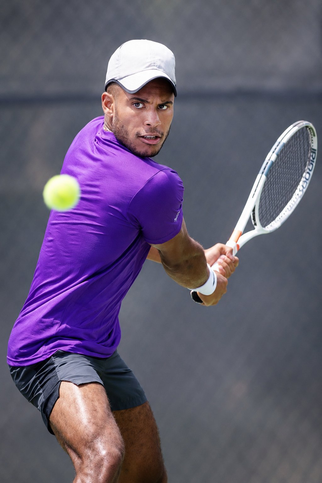 Male tennis player in purple shirt and white cap hitting a forehand shot with a tennis ball and racket on court