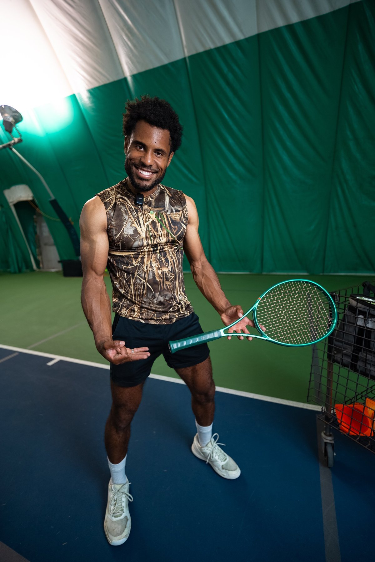 Man holding tennis racket on indoor court, wearing patterned sleeveless shirt and white sneakers