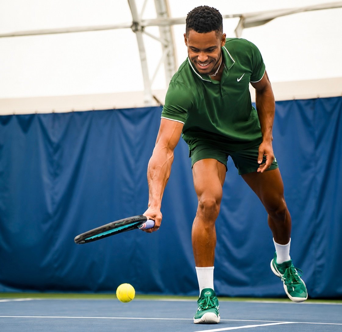 Male tennis player in green shirt preparing to hit a yellow ball on indoor court with blue backdrop