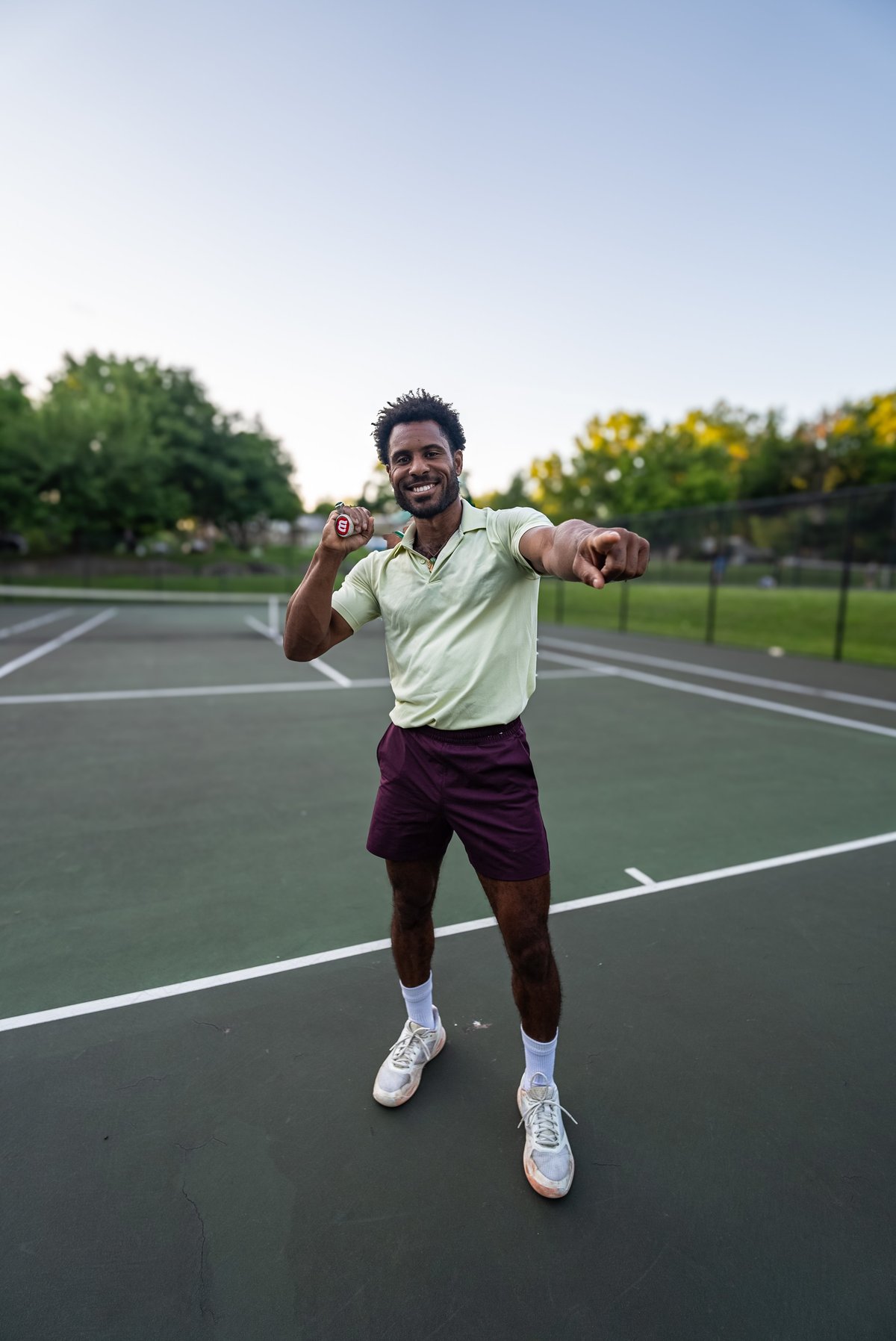 Man in light green shirt and purple shorts celebrates on tennis court with trees in background