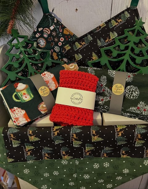Christmas gift wrapping display with festive paper, green tree cutouts, red knitted item, and holiday-themed fabric on white wooden background