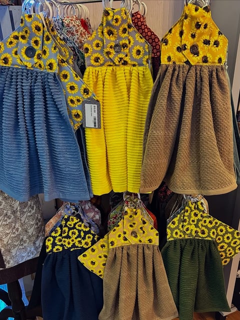 Six colorful childrens dresses on hangers, featuring yellow sunflower print tops and solid-colored pleated skirts in blue, yellow, beige, black, and green.