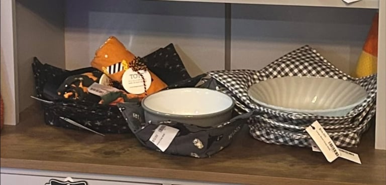 Display of Halloween-themed tableware and decorations including orange pumpkin, black checkered pattern items, white bowls, and festive skull design on wooden shelf