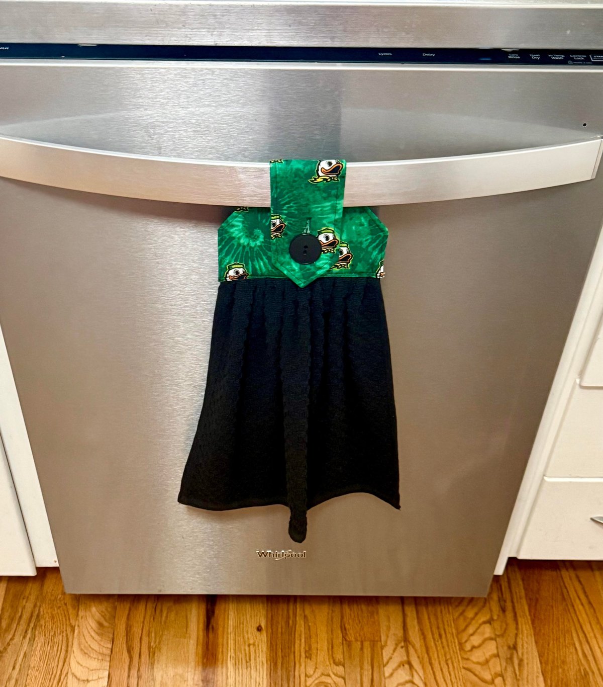 A decorative kitchen towel with a green patterned top and black fabric hanging from a stainless steel dishwasher handle