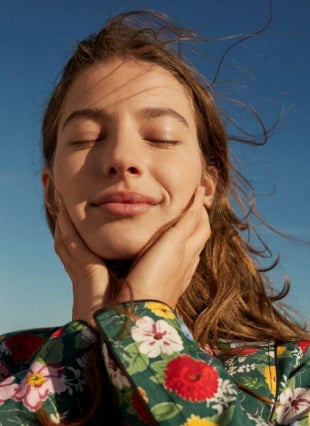 Woman with long brown hair in a floral print shirt, eyes closed, touching her face against a blue sky background