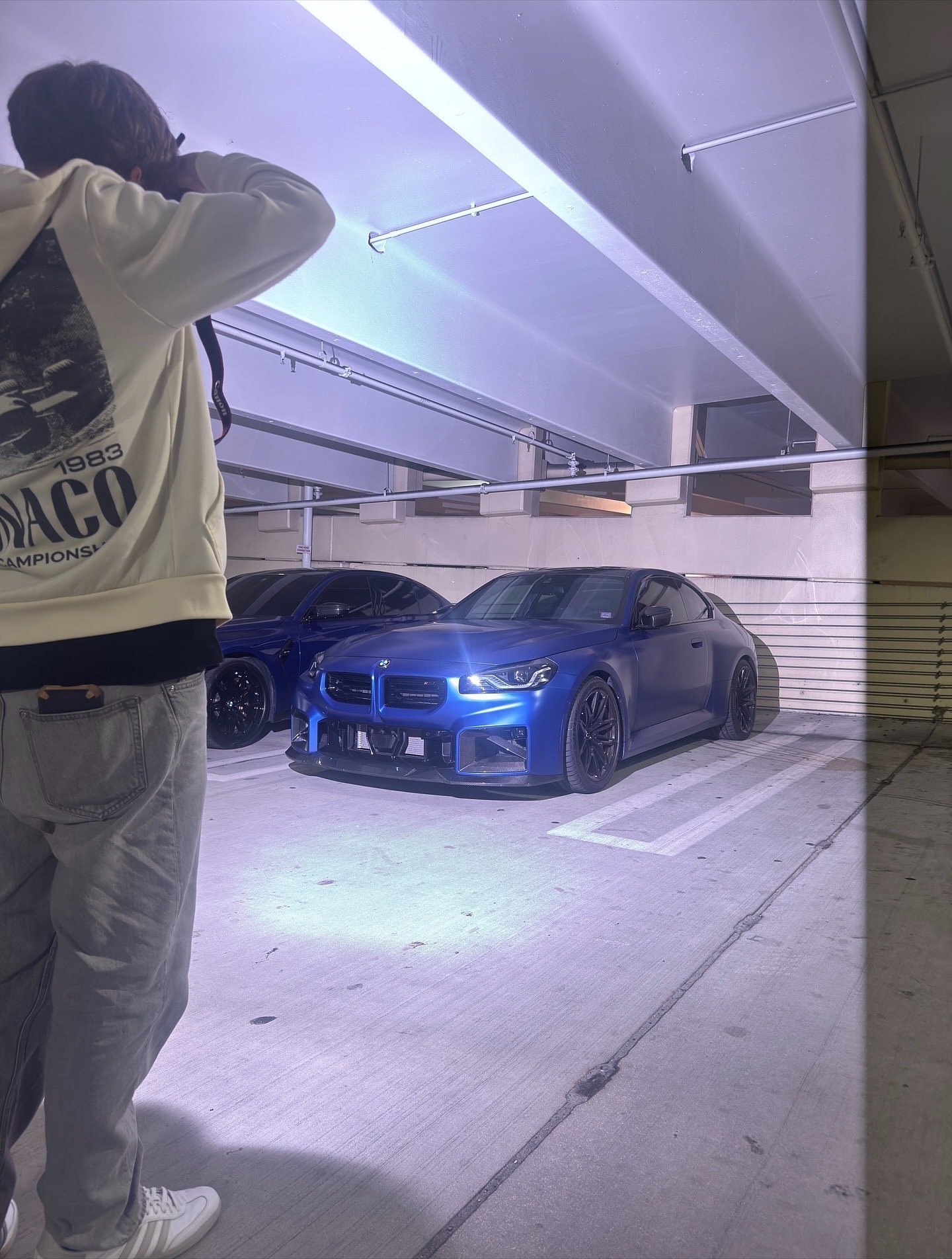 Person photographing a blue sports car in an underground parking garage with purple lighting