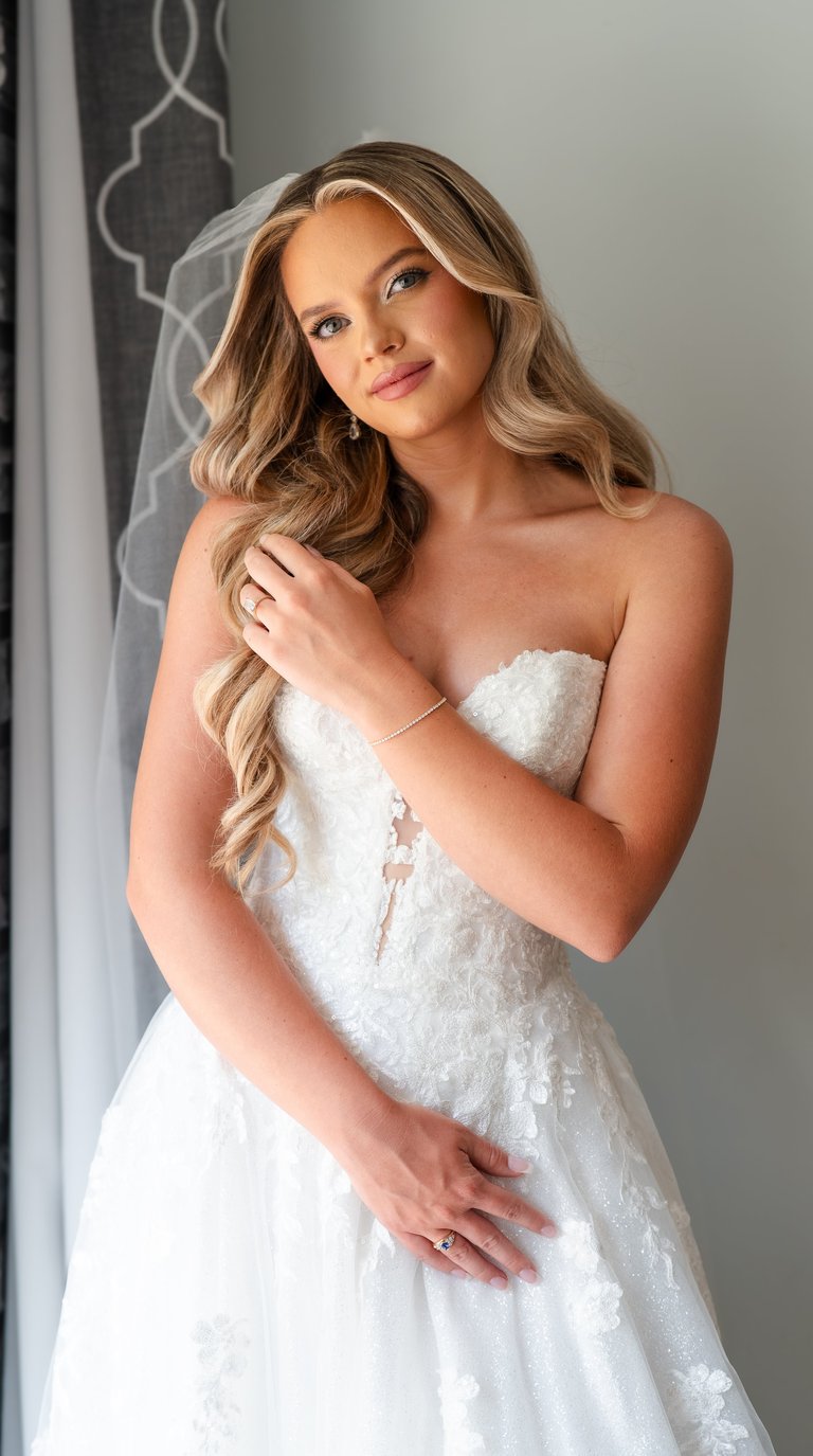 Woman with long wavy blonde hair wearing an ornate white strapless wedding dress, posing with hand in hair against a neutral background