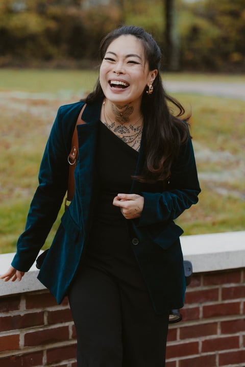 A woman laughing outdoors wearing a dark blue blazer and black pants, with a red and yellow bag strap visible, standing in front of a brick wall and blurred park background
