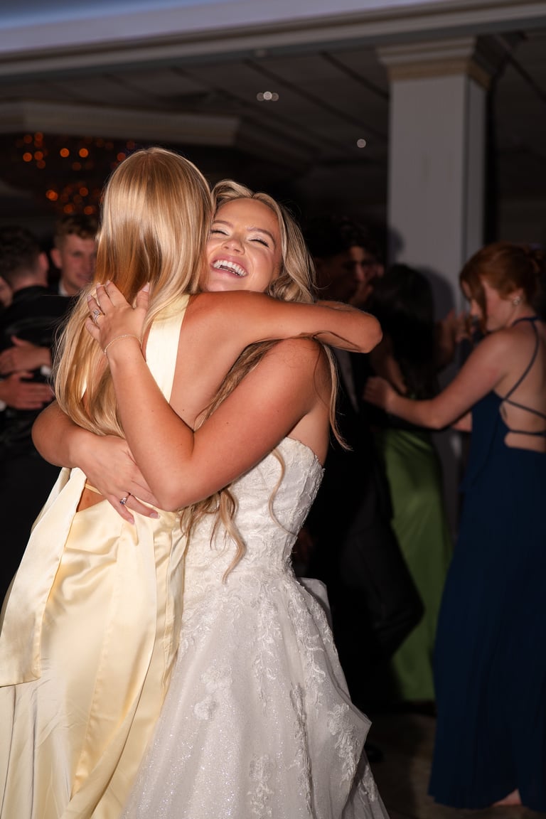 Two women embracing warmly at an indoor event, one wearing a white dress and another in a pale yellow dress, with guests visible in the background