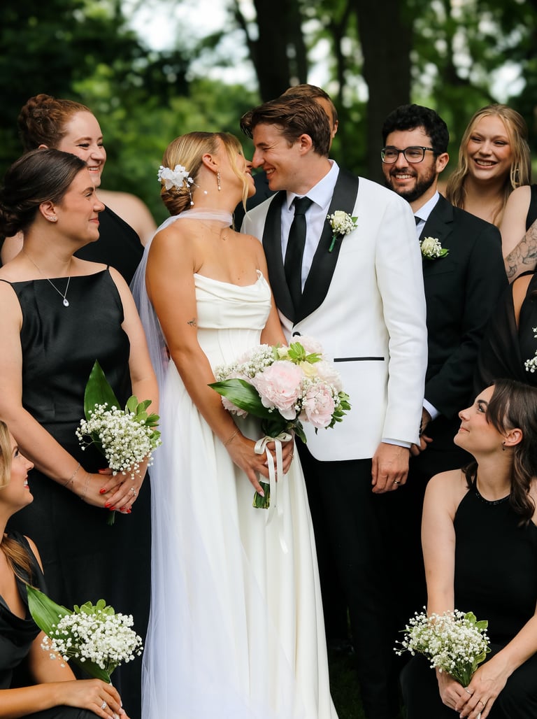 Bride in white dress and groom in light jacket surrounded by wedding party holding white flowers outdoors