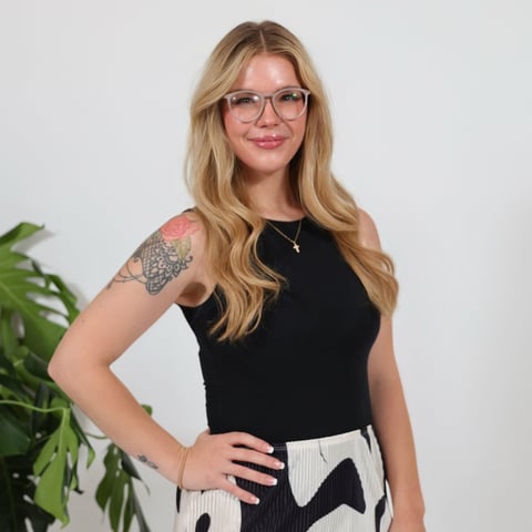 Portrait of a woman with blonde wavy hair and glasses wearing a black sleeveless top, with a visible tattoo on her shoulder and a plant in the background