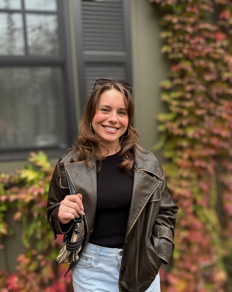Young woman smiling at camera wearing brown leather jacket and black top in front of ivy-covered wall