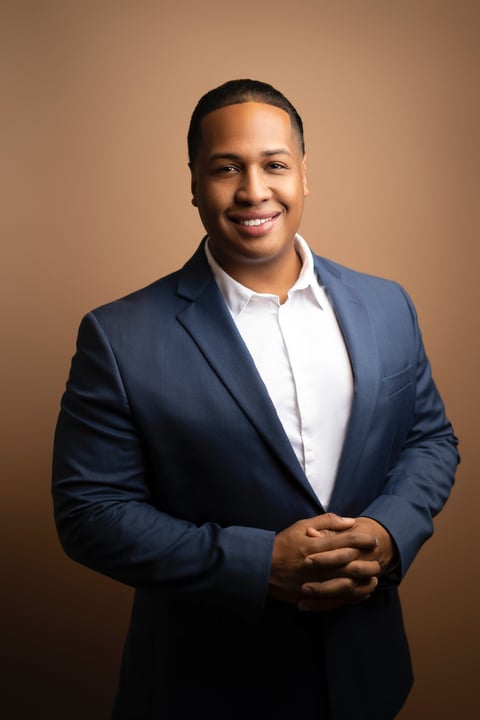Professional portrait of a man in a navy blazer and white shirt smiling against a tan background