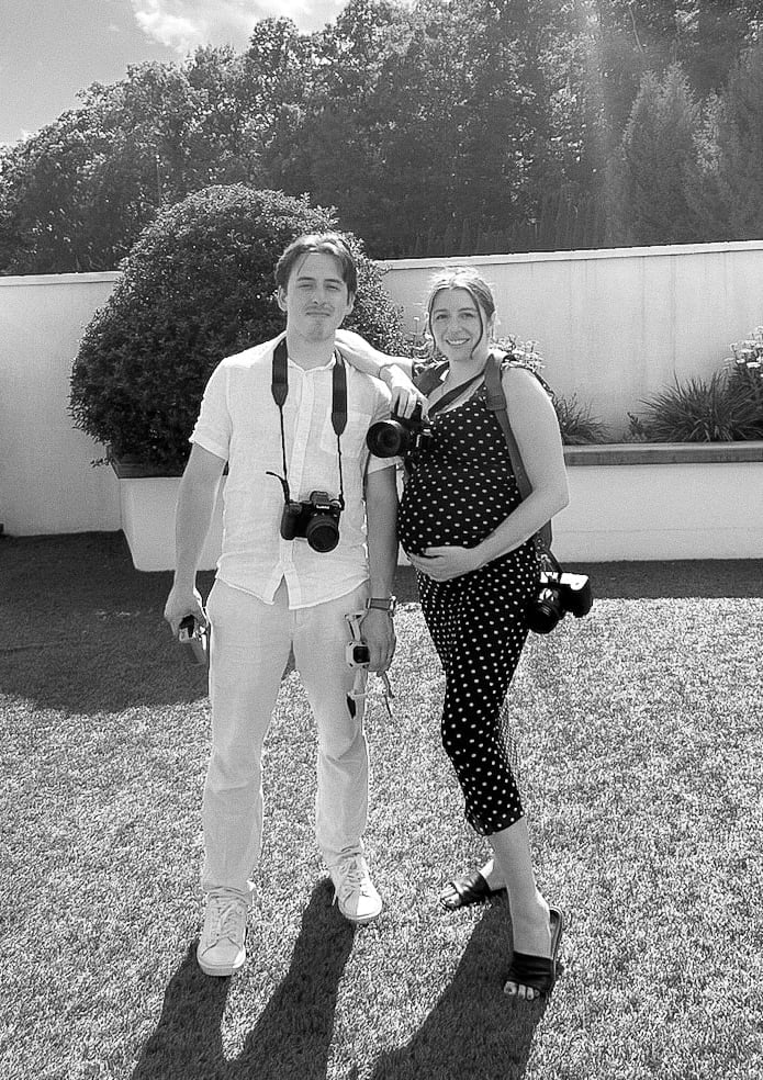 Black and white photo of a man and woman posing outdoors. Man wears light suit with camera, woman wears polka dot top. Modern building and landscaping in background.