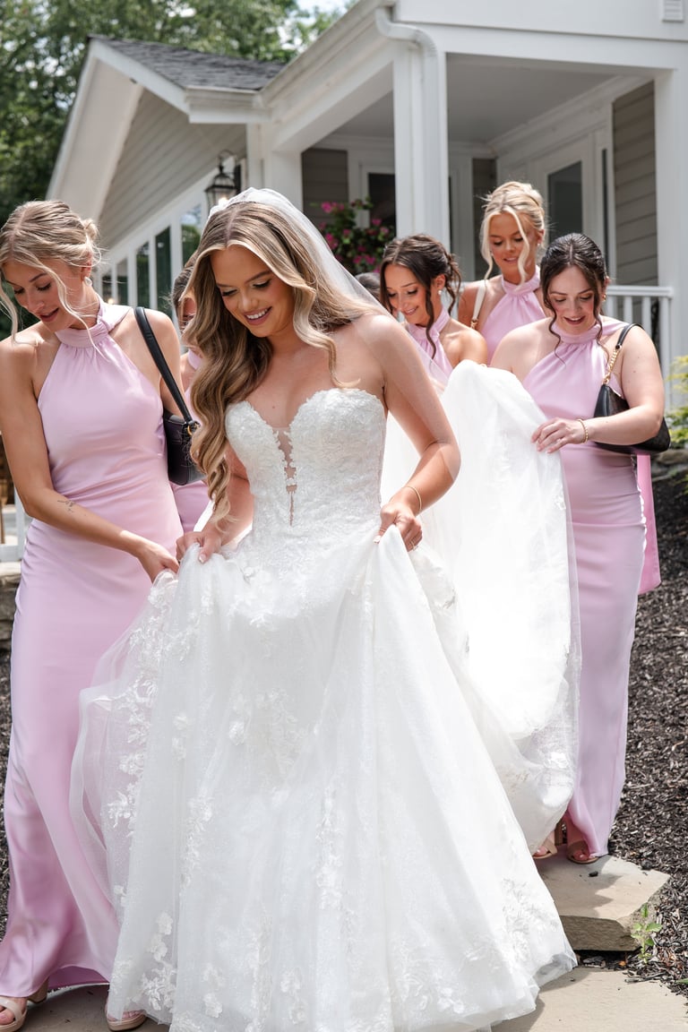 Bride in white lace wedding dress standing with bridesmaids in pink dresses outside a white house