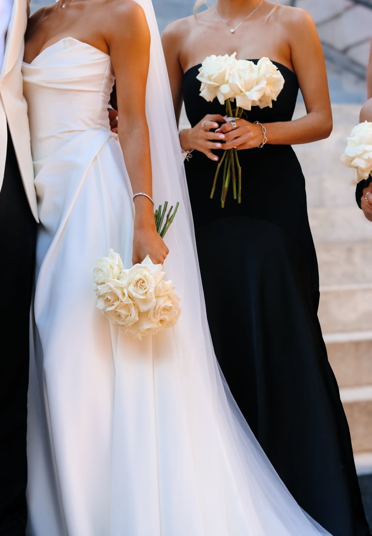 Bride in white strapless dress holding cream roses bouquet standing next to bridesmaid in black strapless dress with white flower bouquet