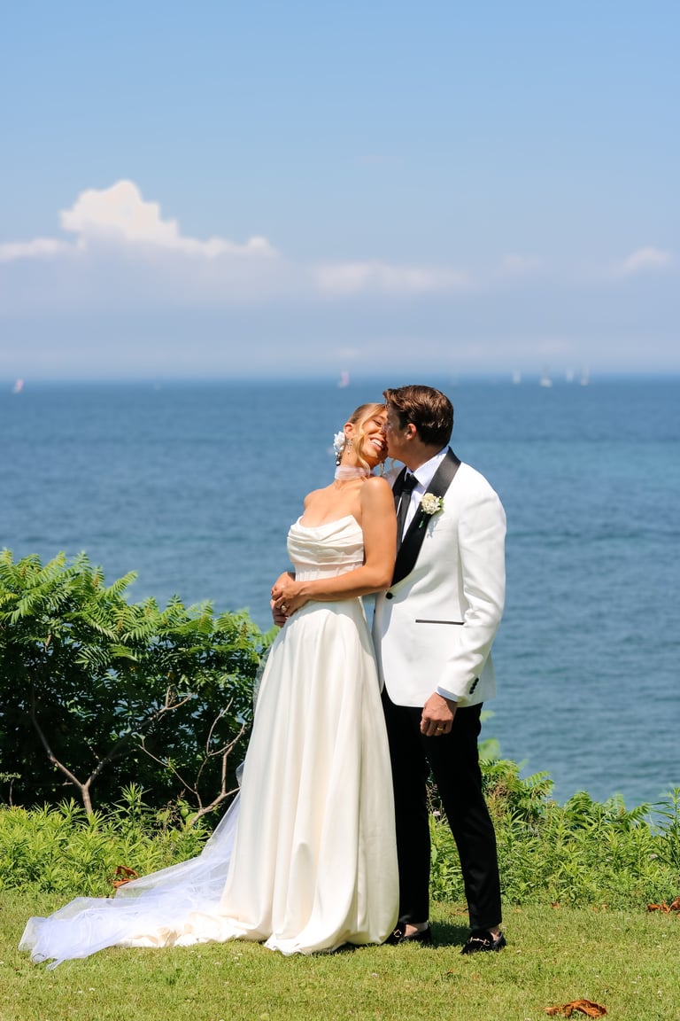Bride and groom embracing on grassy clifftop overlooking ocean on their wedding day