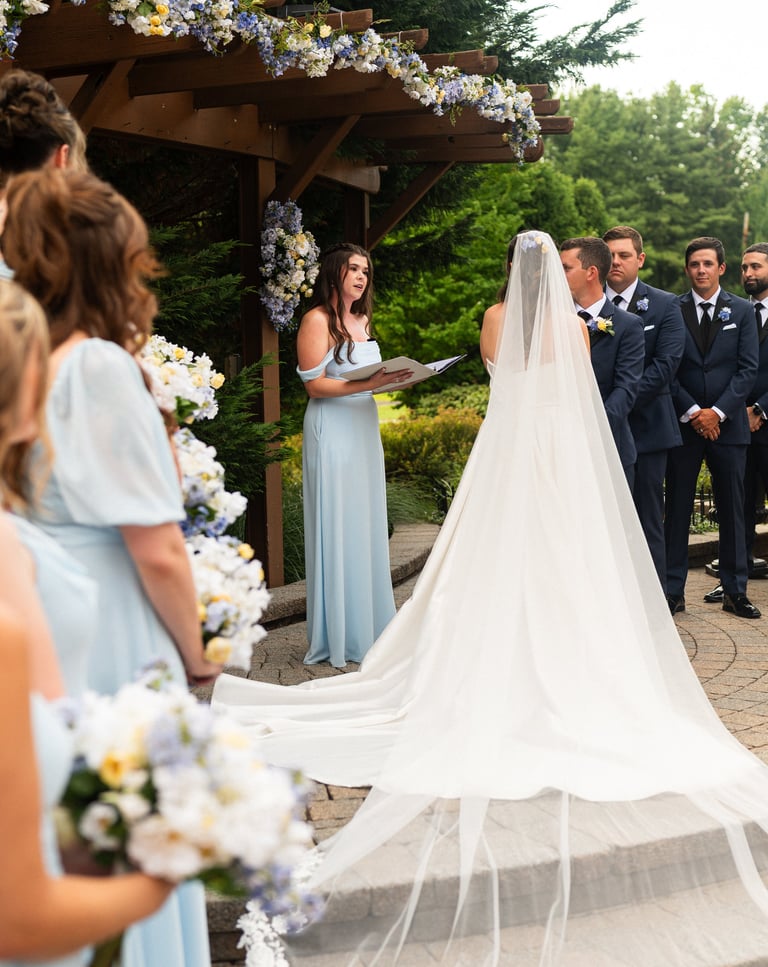 Outdoor wedding ceremony with bride in white dress and veil at wooden arbor decorated with white flowers, groom in navy suit, and wedding party