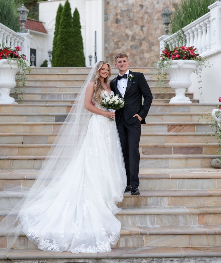 Bride and groom posing on stone steps outside elegant venue, bride in white lace gown with veil holding bouquet, groom in black tuxedo