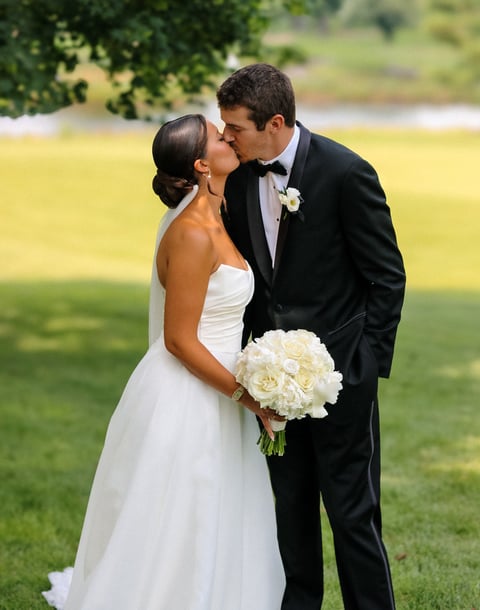 Bride and groom kissing outdoors on lawn, bride in white strapless gown holding white flower bouquet, groom in black tuxedo with boutonniere