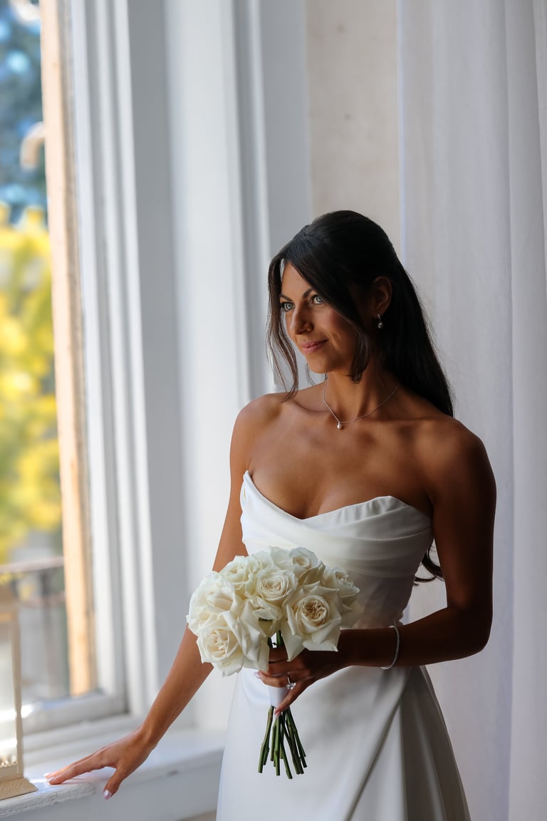 Bride in white strapless wedding dress holding white rose bouquet, standing by bright window with white curtains