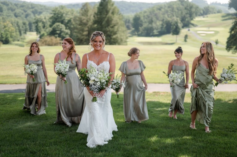 Bride in white wedding dress stands with bridesmaids in gray dresses holding bouquets on a green lawn with scenic valley background
