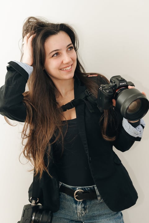 Woman photographer with long dark hair holding a Sony camera with telephoto lens, wearing black blazer and denim, smiling at camera