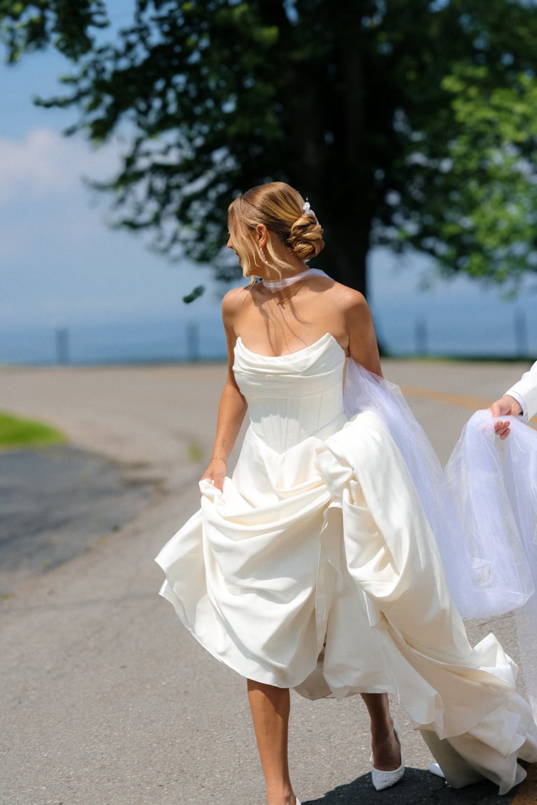 Bride in white wedding dress walking outdoors on a sunny day near water and trees, viewed from behind