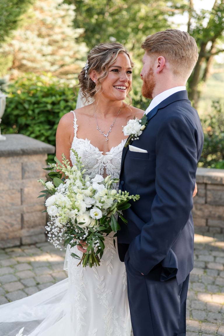 Bride and groom smiling at each other, holding white floral bouquet in garden setting
