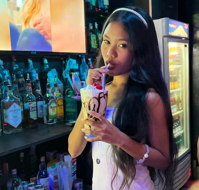 Woman with long black hair and white headband sipping a colorful drink in front of bar shelves with bottles
