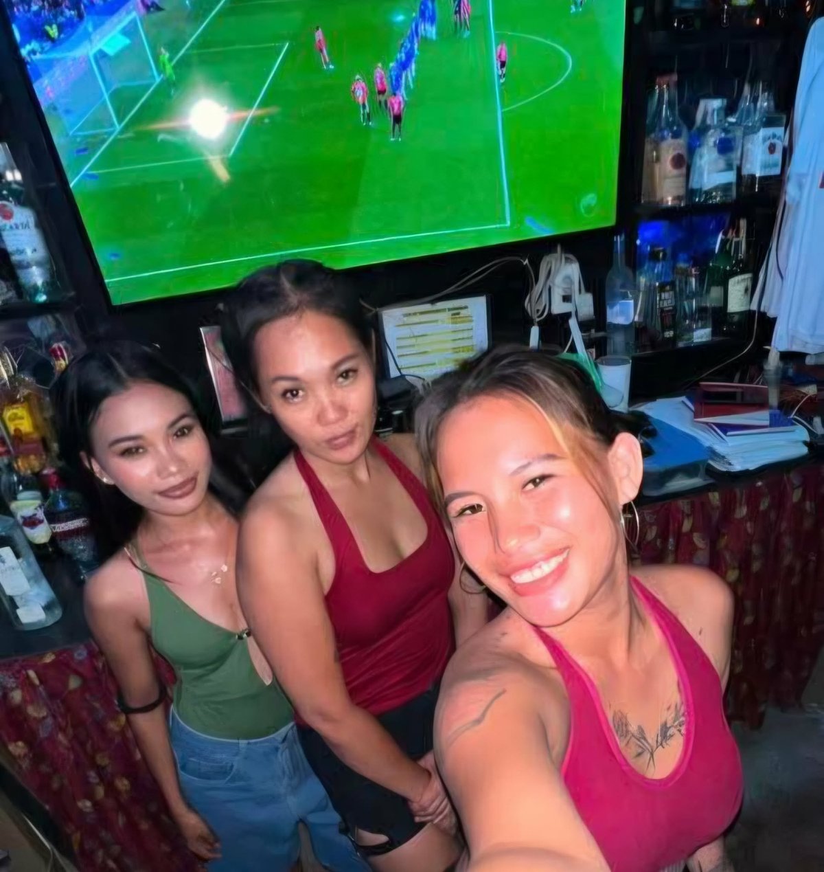 Three women taking a selfie at a bar with a soccer match displayed on a TV screen behind them